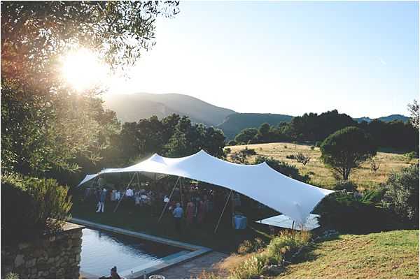 Wide aerial-style shot of an outdoor wedding reception taking place beneath a large white stretch tent, positioned alongside a rectangular pool in a rural hillside setting. A crowd of approximately 30–50 guests mingles under the tent at what appears to be a cocktail hour or reception, with the image taken during golden hour as the sun flares low on the horizon. The surrounding landscape features open fields and rolling mountains in the background. Potential venue feature image.