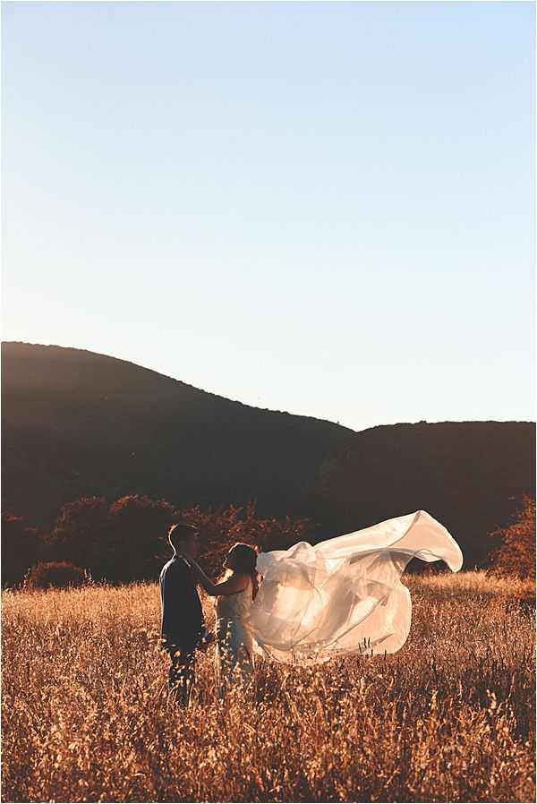 A couple portrait taken outdoors in a dry golden field at sunset, with rolling hills silhouetted in the background. The bride wears a white gown with a long veil or train that billows dramatically in the wind, extending several meters to the right of the frame. The groom is dressed in a dark suit and faces the bride, holding her hands. The warm golden-hour light bathes the scene, creating a strong contrast between the glowing field and the dark hillside backdrop. Wide shot composition with the couple positioned in the lower left third of the frame.