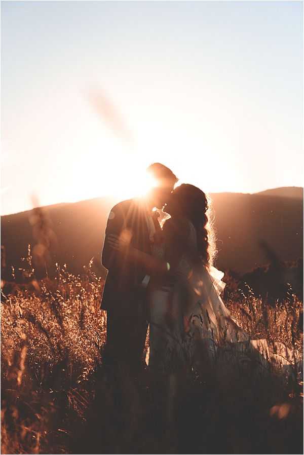 A couple shares a kiss during an outdoor portrait session in a wild grass field at golden hour, with the setting sun positioned directly behind them creating a strong backlit silhouette effect and visible lens flare. The warm orange and amber tones of the sunset illuminate the dried grasses in the foreground and rolling hills in the background. The bride wears a long flowing dress with a veil, and her hair falls in loose waves over her shoulder; the groom is in a dark suit. The composition is a medium-wide portrait shot with the couple centered against the bright sun, giving the image a warm, high-contrast, backlit aesthetic suited to a boho or natural outdoor wedding style.