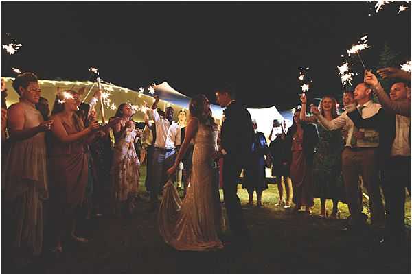 A sparkler send-off exit scene photographed outdoors at night, with the bride and groom sharing a kiss at the center of a corridor formed by approximately 20 guests holding lit sparklers on either side. The bride wears a fitted, beaded or sequined ivory gown and the groom is in a dark suit. Guests are dressed in a mix of casual-to-semi-formal attire, including a dusty mauve dress visible on the left side. In the background, a large white sailcloth or stretch tent is warmly lit, serving as the reception marquee. The sparklers provide the primary light source, creating a warm golden glow against the dark night sky. Wide-shot composition captures the full length of the sparkler tunnel and surrounding guests.