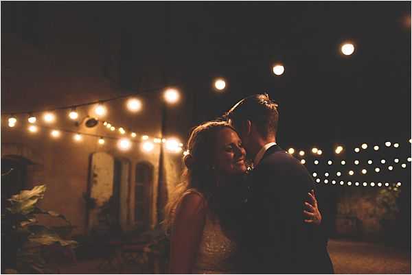 A couple shares a close romantic moment outdoors at night, likely during an evening reception. The bride wears a sleeveless dress with what appears to be a beaded or lace bodice, and the groom is in a dark navy suit, with his back to the camera. The setting is an outdoor courtyard or terrace against a warm stone building, strung with globe fairy lights on overhead cables that create a warm amber glow across the scene. The image is a close portrait with moody, low-light exposure, the couple lit primarily by the warm string lighting against a dark night sky.