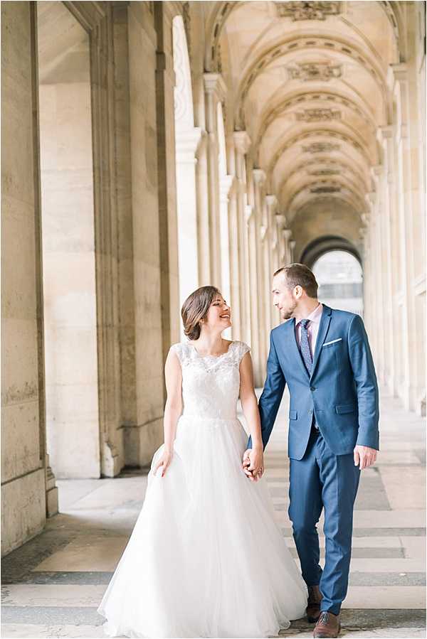 A couple portrait taken beneath a grand arched colonnade, likely at the Louvre or Palais Royal in Paris, featuring a long vaulted gallery with ornate painted ceiling details and tall stone columns receding into the background. The bride wears an ivory A-line gown with a lace cap-sleeve bodice and full tulle skirt, while the groom wears a fitted navy blue suit with a mauve patterned tie and white pocket square. The two are walking hand-in-hand, laughing and looking at each other, captured in a candid mid-stride pose. The composition is a medium full-length portrait shot with the colonnade providing strong leading lines toward a soft background.