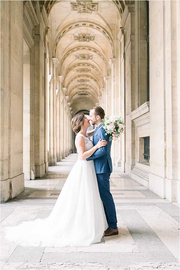 A couple portrait taken beneath the ornate vaulted arcades of a grand Parisian neoclassical building, likely the Louvre or Palais Royal, featuring repeated arched ceilings with decorative carved details receding into the background. The bride wears a white sleeveless ballgown with a lace bodice and full tulle skirt with a train, while the groom wears a navy blue suit with brown dress shoes. The couple is nearly kissing, with the groom's hand on her face and the bride holding a loose, garden-style bouquet of blush pink roses, white blooms, and greenery. The shot is a full-length portrait framed symmetrically within the arcade colonnade, taken at ground level with a shallow depth of field that keeps the couple sharp against the softly blurred architectural corridor. Potential venue feature image.