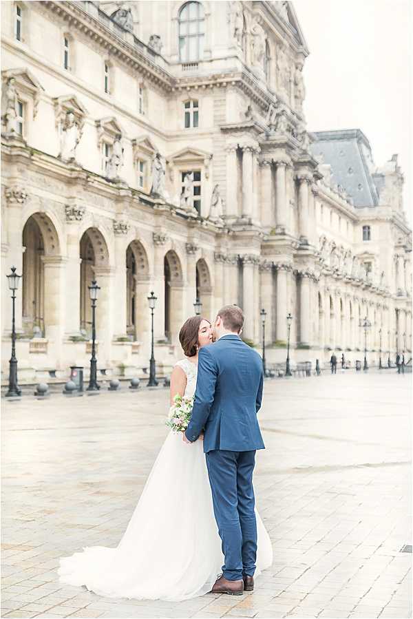 A couple shares a kiss during an outdoor portrait session in the courtyard of the Louvre museum in Paris, with the ornate Baroque facade stretching across the background. The bride wears a white ball gown with a lace bodice and a full tulle skirt with a trailing train, and holds a small bouquet with soft pink and white blooms. The groom wears a navy blue suit with brown dress shoes. The image is a medium-distance portrait shot from behind, with the couple centered against the pale stone architecture and classic black lampposts lining the cobblestone courtyard.