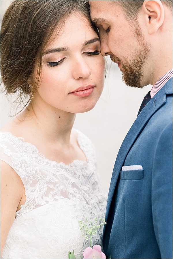A close-up portrait of a bride and groom with their foreheads touching and eyes closed in an intimate pose. The bride wears a white lace dress with a scalloped neckline and cap sleeves, her dark hair styled in a loose updo, and holds what appears to be a light pink tulip with small greenery. The groom wears a navy blue suit with a pink checked shirt and a dark polka-dot tie, with a light blue pocket square. The background is soft and neutral, consistent with a light-toned wall or backdrop, giving the image an airy, fine-art feel.