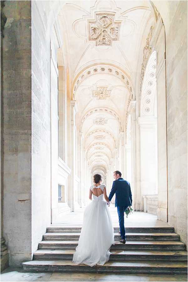 A couple portrait shot taken from behind as the bride and groom walk hand-in-hand up a wide stone staircase beneath a grand classical arcade with ornate plasterwork vaulted ceilings and repeating arched columns receding into the distance. The bride wears a white ballgown with a lace cap-sleeve bodice featuring a keyhole open back and a full trailing skirt, while the groom is dressed in a navy suit; he carries a small green foliage bouquet. The wide shot uses the strong architectural perspective of the colonnade as a leading line, creating depth and framing the couple centrally in the composition. Potential venue feature image.