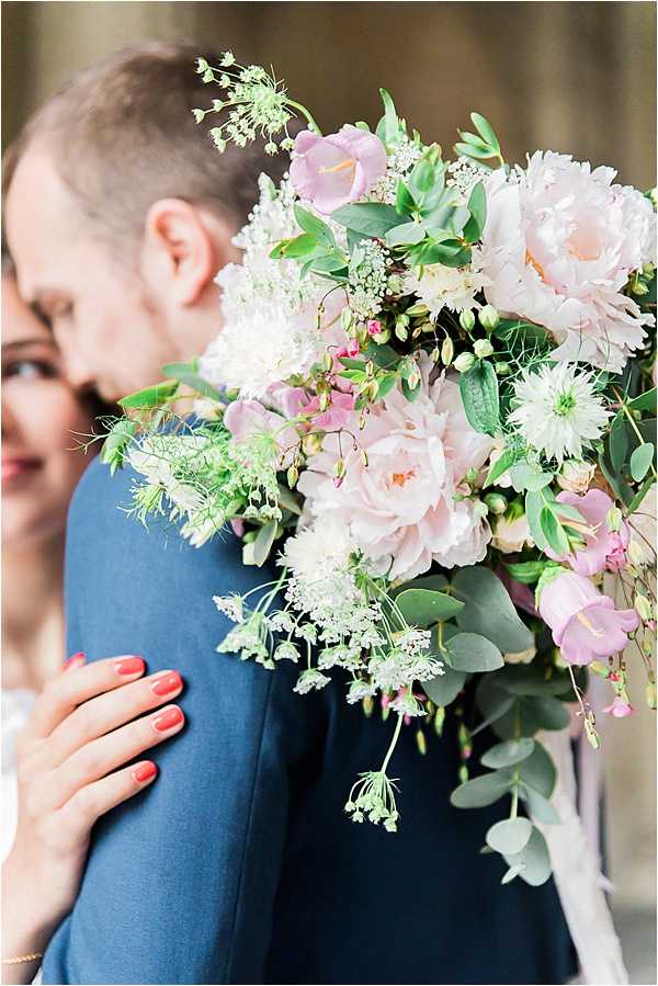 A close-up portrait of a bride embracing a groom from behind, with the bouquet prominently featured in the foreground. The groom wears a navy blue suit jacket, and the bride's hand — with coral-red nails — rests on his shoulder. The bridal bouquet is a loosely arranged garden-style arrangement featuring blush peonies, pale pink roses, mauve campanula bells, white scabiosa, queen anne's lace, and eucalyptus foliage with trailing greenery. The composition is a tight detail shot with the couple softly out of focus in the background, drawing full attention to the bouquet's texture and color palette of blush, mauve, and green.