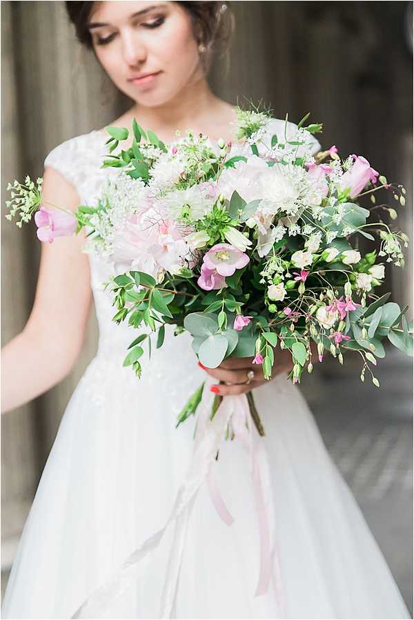 A close-up portrait of a bride looking down at her bouquet, set against a softly blurred architectural background. She wears a white cap-sleeve lace bodice dress with a tulle skirt, with her dark hair pulled up. The bouquet is a loose, garden-style arrangement featuring blush peonies, pink lisianthus, white Queen Anne's lace, cream freesia buds, eucalyptus, and trailing greenery, tied with a long blush pink ribbon. The composition is a medium portrait shot with the bouquet in sharp focus in the foreground and the bride's face softly focused in the background.
