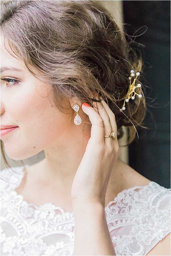 Close-up portrait of a bride adjusting her earring during the getting-ready portion of the day. She wears gold teardrop crystal drop earrings and a gold ring with a clear stone, and her hair is styled in a loose updo adorned with a gold and pearl hair pin. Her white lace gown features a cap sleeve with detailed floral lace appliqué. Her nails are painted coral-red, and she wears natural, soft bridal makeup.