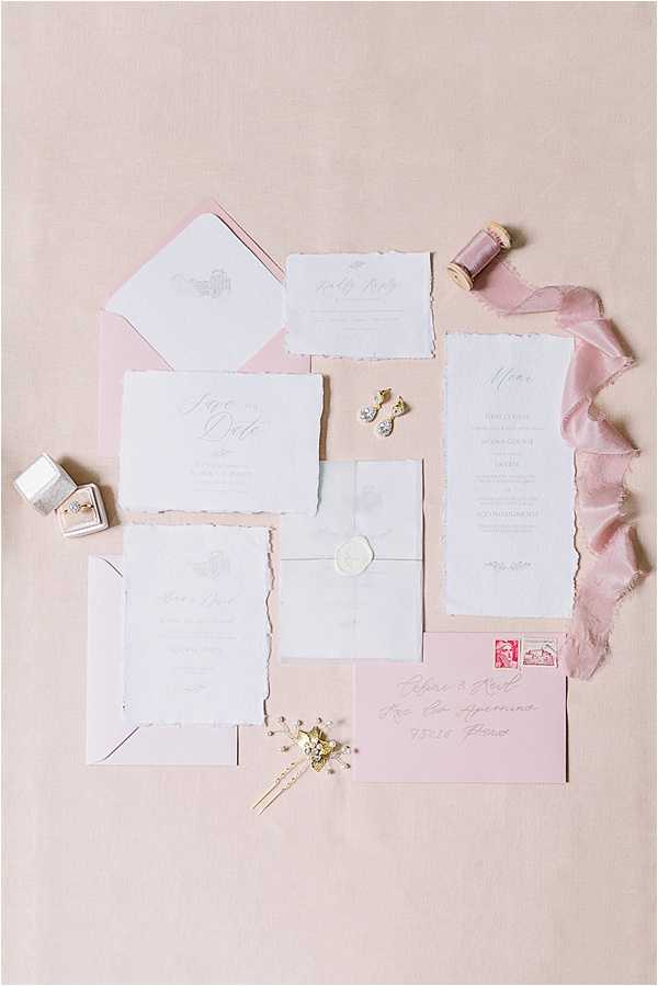 A flat lay photograph of a wedding stationery suite arranged on a blush pink linen surface. The suite includes deckle-edged paper cards in white and pale grey featuring a save the date, RSVP card, menu card, invitation, and a vellum envelope sealed with a white wax seal, alongside blush pink envelopes — one addressed with calligraphy script and bearing a red French postage stamp. Styled accessories include a small velvet ring box in light grey holding a diamond ring, a pair of gold drop earrings with pearl and crystal details, a gold floral hair pin, and a spool of mauve-toned silk ribbon with frayed chiffon ribbon in blush pink draped across the top right corner. The overall palette is blush, white, and pale grey with gold accent details, reflecting a romantic, classic styling approach.