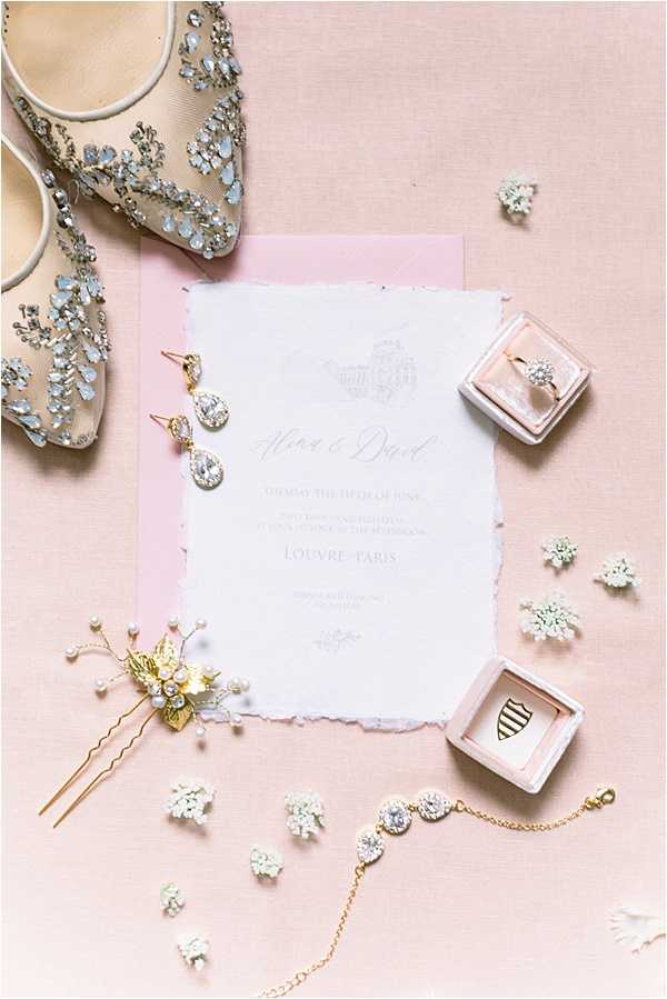 A flat lay detail shot of a bridal accessories and stationery arrangement on a blush pink linen background. The centerpiece is a white deckle-edged wedding invitation with script typography referencing the Louvre, Paris, placed on a blush pink envelope. Surrounding the invitation are crystal drop earrings, a gold and pearl hair pin with floral detailing, a gold chain bracelet with round crystal links, two small blush velvet ring boxes (one containing a diamond solitaire ring, one containing a crest-engraved piece), and a pair of nude mesh heels heavily embellished with silver and crystal beading. Small sprigs of white dried florals are scattered throughout the composition as decorative accents.