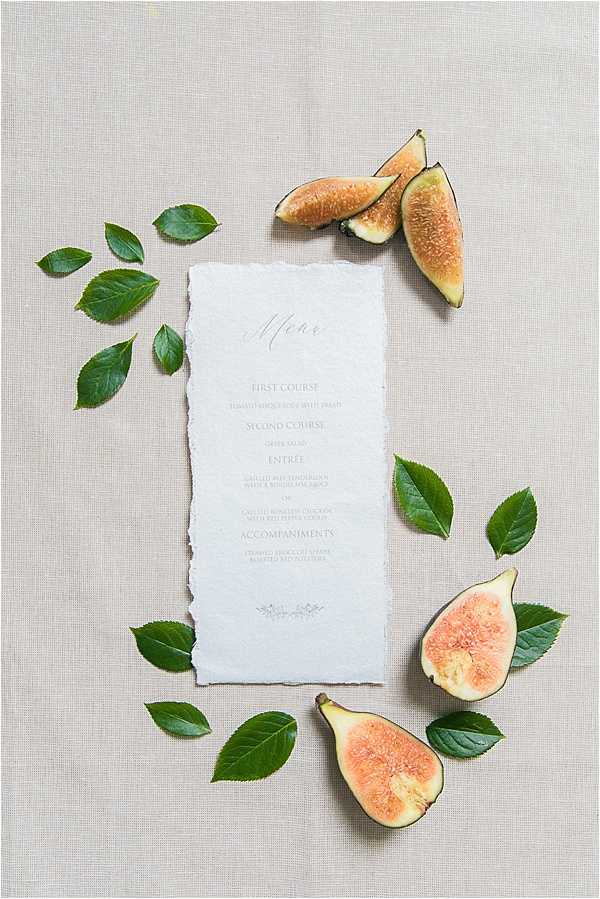 A flat lay detail shot of a wedding menu card printed on white deckle-edged paper with grey serif and script typography, listing courses including First Course, Second Course, Entrée, and Accompaniments. The menu is styled on a neutral linen-textured surface and surrounded by scattered dark green leaves and freshly cut figs showing their orange-pink interior flesh, arranged decoratively around the card's perimeter. The overall styling palette is soft and neutral with natural organic accents, reflecting a refined rustic or garden-inspired aesthetic.