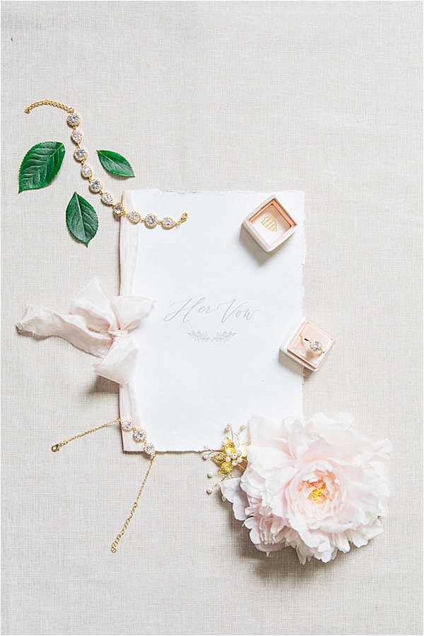 A flat lay detail shot of bridal accessories arranged on a neutral linen-textured surface. The central piece is a white deckle-edge vow booklet with calligraphy reading 'Her Vows,' layered over a slightly larger white paper with a blush pink silk ribbon. Two gold and crystal diamond-style bracelets are placed around the stationery, along with a blush velvet ring box containing a solitaire diamond engagement ring and a second closed blush velvet ring box with gold monogram detailing. A large blush pink peony bloom sits at the lower right, accompanied by small gold and pearl hair pins, while a few loose green leaves accent the upper left corner. The overall palette is blush pink, ivory, and gold with a soft, classic bridal styling theme.