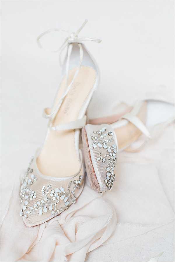 A close-up detail shot of a pair of bridal heels placed on a soft blush fabric, shot against a light neutral background. The shoes feature embellished toe caps with silver crystal and rhinestone floral embroidery on a blush mesh base, paired with ivory satin ankle-tie straps and a block heel. The insole is visible on one shoe, showing a designer label. The overall styling palette is blush and ivory, consistent with a classic or romantic bridal aesthetic.