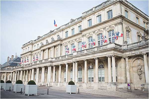 Wide exterior shot of a large neoclassical French government or civic building featuring a colonnade of tall stone columns along the ground floor, ornate stone facade, and multiple balconies decorated with French tricolor flags. The building appears to be the Hôtel de Ville or a préfecture, likely in Bordeaux. No people, wedding party, or decor are visible in the frame. Potential venue feature image.