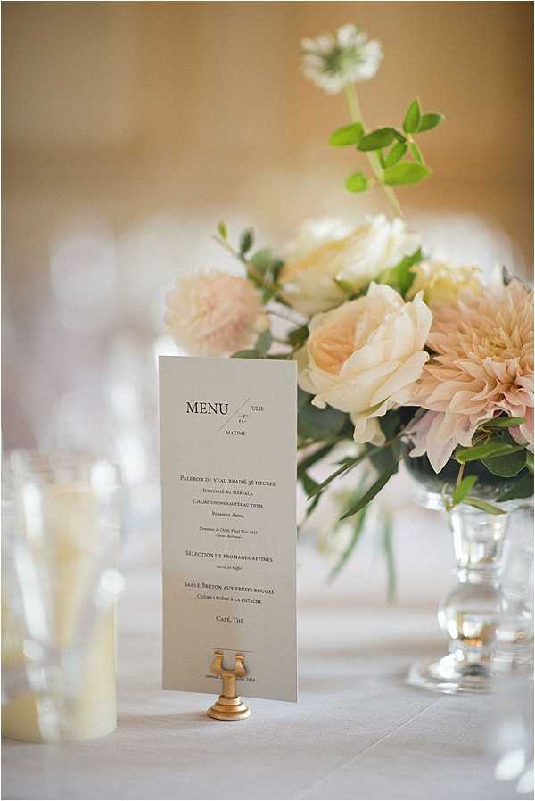 A close-up detail shot of a wedding reception table setting featuring a printed French menu card for Julie and Maxime, displayed in a small gold brass holder on a white linen tablecloth. The menu lists several courses including braised veal, cheese selection, and a Breton sablé dessert. Behind the menu card sits a low floral centerpiece in a clear glass footed vase containing blush dahlias, cream garden roses, soft pink pom-style blooms, and eucalyptus greenery. A white pillar candle in a glass holder is partially visible to the left. The overall decor palette is blush, ivory, and gold with a classic, refined French reception aesthetic.