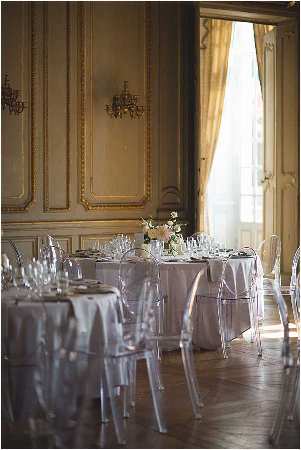 Indoor wedding reception setup inside a French chateau ballroom featuring ornate gold-trimmed boiserie wall paneling, wrought iron wall sconces, and parquet flooring. Round tables are dressed in pale grey-white linen with clear acrylic ghost chairs, crystal glassware, and small low centerpieces of blush pink and white flowers including what appear to be peonies and daisies. Natural light streams through tall floor-to-ceiling doors dressed with gold-toned curtains. The overall decor palette is soft grey, white, and blush with a classic French interior aesthetic. Wide shot capturing the full room setup with no guests present. Potential venue feature image.