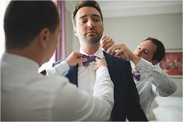 A getting-ready scene showing a groom in a navy blue suit jacket and white dress shirt having a purple bow tie adjusted by two groomsmen, both dressed in white shirts. The three men are indoors, likely in a hotel room, with a bed and light-colored walls visible in the background. The groom stands in the center looking downward while one groomsman adjusts the bow tie from the front and another assists from the right side. The shot is a mid-range portrait with a shallow depth of field, slightly blurring the background and the groomsman in the foreground.