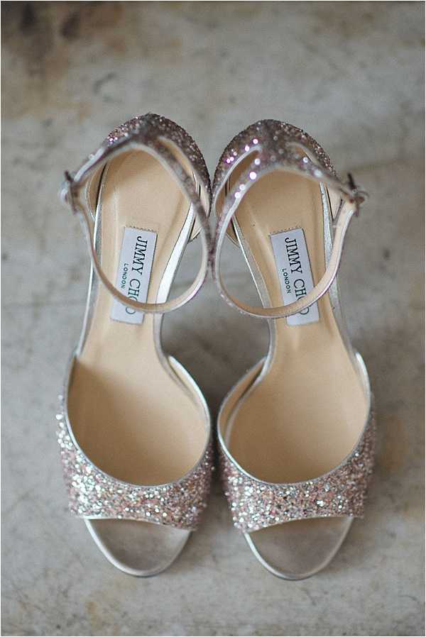 Close-up flat lay detail shot of a pair of Jimmy Choo London bridal sandals placed sole-up on a stone surface. The shoes feature open-toe strappy silhouettes with rose gold and silver glitter-covered toe straps and heel counters, thin ankle strap buckle closures, and nude/champagne leather insoles with the Jimmy Choo London brand labels visible. The glitter detailing catches light across both shoes, and the overall color palette is rose gold, silver, and champagne.