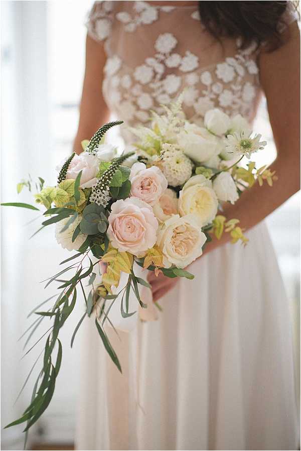 Close-up portrait of a bride holding a loose, cascading bridal bouquet composed of blush garden roses, cream David Austin roses, white ranunculus, white astilbe, scabiosa, green cymbidium orchids, veronicastrum, eucalyptus, and trailing willow-like greenery. The bride wears a white gown with a sheer lace cap-sleeve bodice featuring floral appliqué over a flowing skirt. The bouquet style is organic and unstructured, with a soft blush, cream, and green palette. The image is a detail-focused portrait shot with a soft, bright background and shallow depth of field.