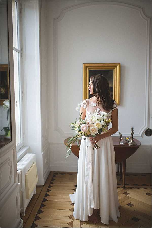 A bride stands alone in a portrait shot inside a classic French interior room, looking toward a tall window with natural light falling across her. She wears a floor-length ivory gown with a sheer illusion neckline and delicate 3D floral appliqué detailing on the bodice and cap sleeves, with a flowing skirt. She holds a loose, garden-style bouquet of blush garden roses, cream peonies, ivory ranunculus, and trailing eucalyptus foliage tied with a dark ribbon. The room features white panelled walls with ornate moulding, a gold-framed oil painting, a dark wood antique side table, a cast iron radiator, and a herringbone parquet wood floor with a geometric dark inlay border. The overall styling is classic and refined with a soft, romantic palette.