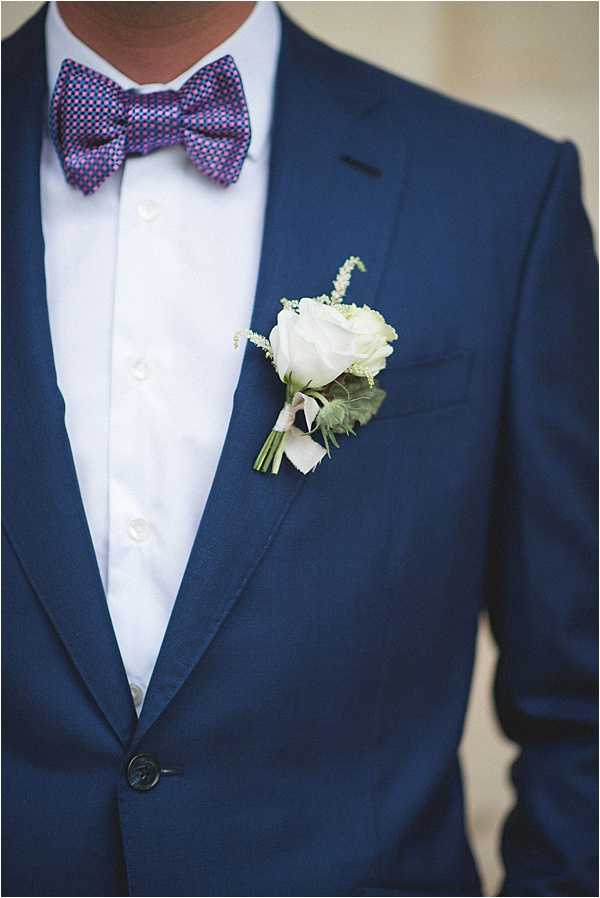 Close-up detail portrait of a groom's torso, showing a navy blue suit jacket worn over a white dress shirt, paired with a purple and pink geometric-patterned bow tie. A boutonniere featuring a single white rose, small white astilbe-like filler flowers, and green foliage is pinned to the left lapel. The composition is a tight crop focusing on the chest and lapel area, with a softly blurred neutral background.