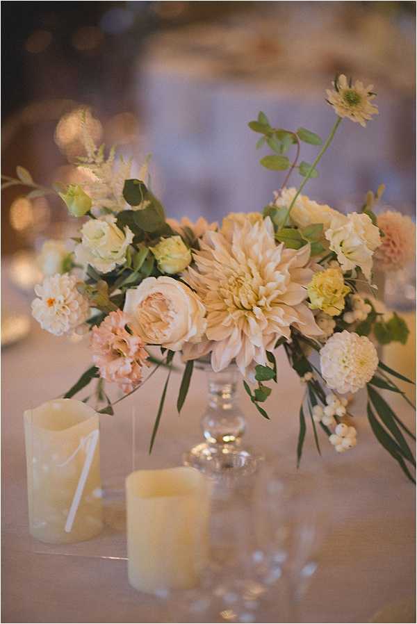 Close-up detail shot of a wedding reception table centerpiece arranged in a clear glass pedestal compote vase. The floral arrangement features large blush and cream dahlias, garden roses, soft yellow button blooms, blush carnations or dianthus, small white berry clusters, and mixed greenery including eucalyptus and palm-like foliage. Two ivory pillar candles are placed in front of the arrangement on a cream or ivory linen tablecloth, along with a small white table number card. The overall decor palette is blush, cream, and soft yellow with a classic, romantic styling. The background shows a softly blurred indoor reception space with warm ambient lighting and white chair covers visible.