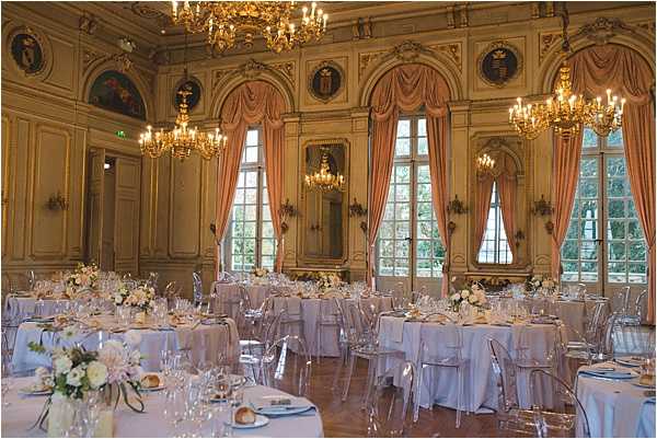 A wide shot of a wedding reception room set up for dinner inside a grand French classical ballroom with gold-panelled walls, ornate painted medallions, and large windows dressed with salmon-pink draped curtains. Multiple round tables are covered in white linens and set with crystal glassware, white china, and low centerpieces of white and blush flowers including what appear to be peonies and roses with greenery. Transparent ghost chairs surround each table. The room is lit by several large gold chandelier candelabras mounted from the ceiling and wall sconces, casting warm amber light throughout the space. The overall decor palette is white, blush, and gold with a classic French formal style. Potential venue feature image.
