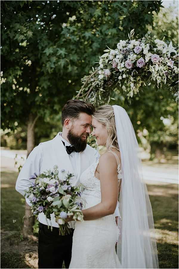 An outdoor couple portrait showing the bride and groom with foreheads touching and smiling at each other in a garden setting. The groom wears a white dress shirt with black bow tie and dark trousers, while the bride wears a fitted lace white gown with spaghetti straps and a long cathedral-length veil. The bride holds a loose, garden-style bouquet of lavender roses, purple anemones, blush blooms, and mixed greenery. Above them, a floral arrangement on a branch or arch features lavender roses, white blooms, and greenery in the same soft purple and white palette. The shot is a medium portrait composition with soft natural daylight filtering through the trees behind them.