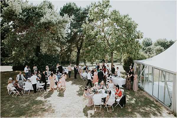 An aerial wide shot of an outdoor cocktail hour or garden reception with approximately 60–80 guests mingling and seated across a shaded lawn. Guests are arranged around small round tables draped in white linens and surrounded by white folding chairs, with a few standing cocktail tables also covered in white cloth. The crowd is dressed in a mix of colorful summer attire including blush, red, and floral dresses. A white marquee tent with glass panels is visible along the right edge of the frame. The setting is an open park-like garden with mature trees providing natural shade over the gathering area.
