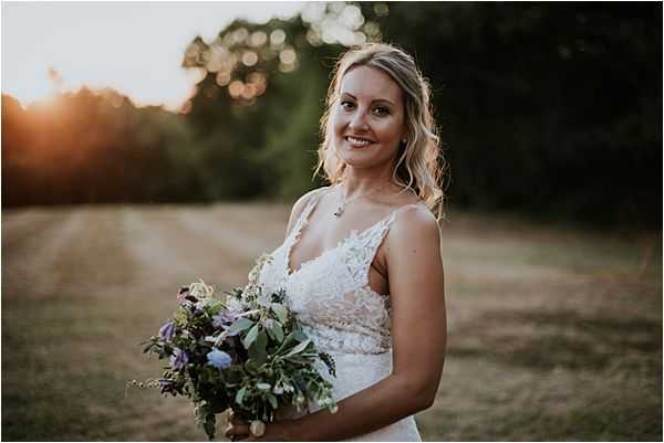 A bridal portrait taken outdoors at golden hour, with warm sunset light glowing behind the bride. The bride wears a sleeveless white lace gown with a V-neckline and spaghetti straps, and holds a loose, garden-style bouquet featuring lavender, light blue hydrangea, purple blooms, and abundant greenery. Her blonde hair is worn down in soft waves. The shot is a close-up portrait with a shallow depth of field, keeping the background softly blurred.