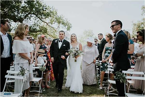 The bride is being walked down the aisle during an outdoor ceremony, escorted by a man in a black tuxedo with a white pocket square on her left and an older woman in a pale pink lace dress and white hat on her right. The bride wears a fitted white lace gown and carries a loose bouquet of purple and white flowers with greenery. Guests line both sides of the grass aisle, seated on white folding chairs decorated with small greenery sprigs, dressed in a mix of casual summer outfits including floral prints and whites. The setting is a garden or parkland area with large trees providing shade, and the ceremony has a relaxed, garden-party aesthetic. Wide shot taken from ground level looking down the aisle toward the approaching processional.