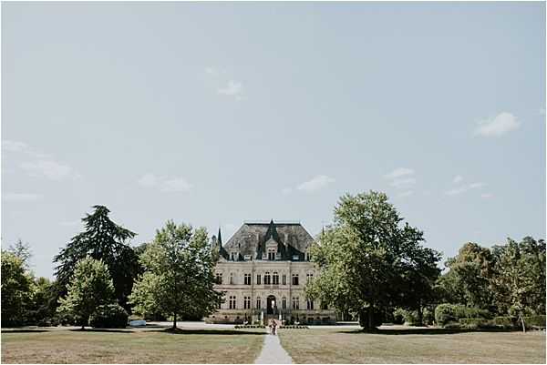 A wide-angle exterior shot of a French chateau with classic 19th-century architecture, featuring a steeply pitched mansard roof, ornate stone facade, and symmetrical window arrangements. Two small figures, appearing to be a couple in wedding attire, stand on the central pathway leading up to the chateau's entrance, providing scale against the grand building. The grounds feature a manicured lawn with a straight gravel or paved approach path flanked by mature trees. Potential venue feature image.