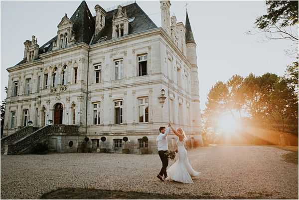 A bride and groom are dancing and spinning on the gravel forecourt of a large French chateau at golden hour, with warm sunset light flaring dramatically from behind the building's right tower. The bride wears a fitted, long white lace gown and holds a bouquet, while the groom is dressed in dark trousers and a white shirt. The chateau is a classic 19th-century French architecture style with pale stone facade, steep slate roofs, ornate dormers, and a corner turret. Wide shot capturing the full building exterior alongside the couple. Potential venue feature image.