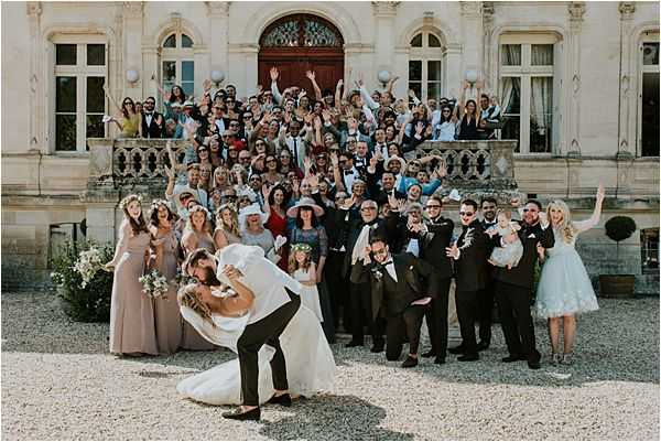 A large group wedding portrait taken outdoors on the gravel forecourt of a French chateau, with the ornate stone facade and grand entrance staircase serving as the backdrop. The groom, dressed in a dark suit, dips the bride in a dramatic kiss at the front of the image while approximately 80-100 cheering guests fan out behind them on the steps and courtyard, many with arms raised. The bride wears a white ball gown and carries a bouquet with white blooms and greenery, while bridesmaids on the left are dressed in blush/taupe floor-length gowns and groomsmen on the right wear dark suits with bow ties. The wide-angle shot captures the full energy of the crowd celebration in what appears to be warm afternoon sunlight. Potential venue feature image.