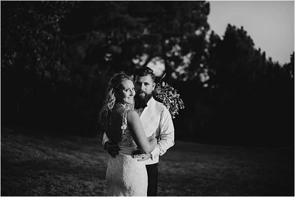 Black-and-white couple portrait taken outdoors at dusk, with dark tree silhouettes in the background creating deep contrast against a lighter sky. The bride wears a fitted lace gown with spaghetti straps and has loose wavy hair, while the groom wears a dress shirt and dark trousers with a full beard; he holds a bouquet of mixed flowers at his side. The couple embraces closely, with the bride turning slightly to look toward the camera and the groom looking slightly away. The image uses moody low-light tones with soft highlights on the subjects and rich dark shadows throughout the background.