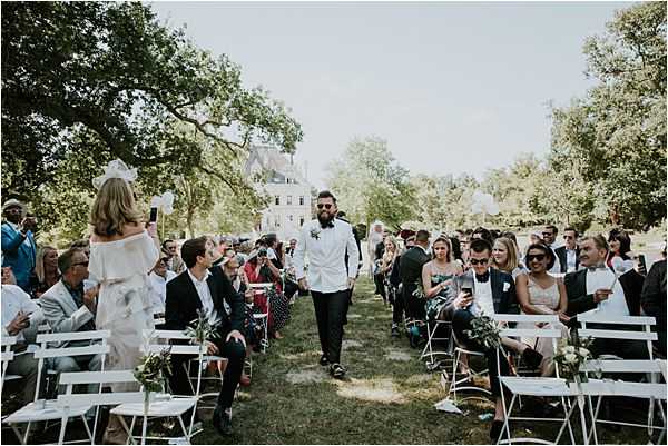 An outdoor wedding ceremony in the grounds of a French chateau, with the groom walking down the aisle toward the altar. The groom wears a white blazer, black trousers, a black bow tie, and sunglasses, with a floral boutonniere. Approximately 40-50 guests are seated on white folding chairs arranged in two rows on either side of a grass aisle, with small greenery arrangements decorating the chair ends. The chateau building with a grey slate roof is visible in the background through the trees. Wide shot taken from behind the guests looking toward the approaching groom, with bright midday sunlight.