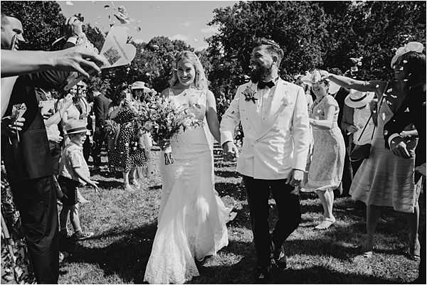 A black-and-white image of a confetti exit following an outdoor wedding ceremony, with the couple walking hand-in-hand through a corridor of approximately 20-30 cheering guests on a grass lawn surrounded by trees. The bride wears a fitted, lace wedding gown with a slight train and carries a full, loosely arranged bouquet, while the groom wears a white double-breasted dinner jacket with dark trousers and a bow tie, with a boutonniere on his lapel. Guests are dressed in summer wedding attire including fascinators, hats, and sundresses, and are tossing confetti from paper cones. The image is a medium wide shot with high contrast black-and-white tones, capturing a candid, joyful moment with strong sunlight creating deep shadows across the scene.