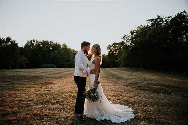 A couple portrait taken outdoors at golden hour in an open field, with the bride and groom facing each other in an intimate pose. The groom wears dark trousers and a white dress shirt with no jacket, while the bride wears a fitted lace wedding dress with a train spread behind her. She holds a loose, organic bouquet featuring deep greenery and what appear to be white and mauve blooms. The warm backlit sunset creates a golden glow around both figures, with a tree line visible in the background. The composition is a medium wide shot with a boho-rustic aesthetic.