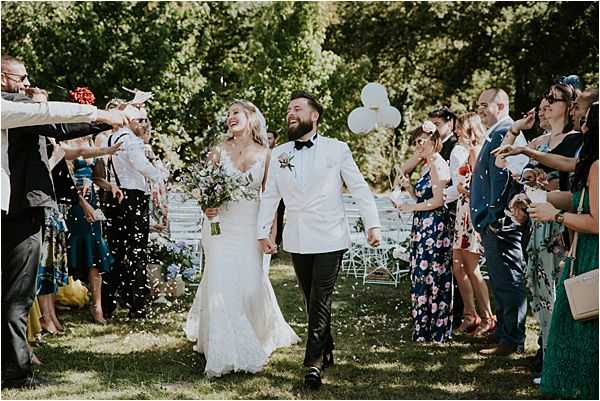 The bride and groom walk back down the aisle during a confetti exit following an outdoor ceremony, with approximately 20-30 guests lining both sides throwing white and pale blue confetti. The ceremony took place on a lawn with white chiavari chairs still visible in the background, along with white balloons. The bride wears a fitted lace wedding gown with a V-neckline and carries a loose, garden-style bouquet featuring blue and white blooms with greenery; the groom wears a white dinner jacket with a black bow tie and dark trousers with a small floral boutonnière. Guests are dressed in colorful summer attire including a floral maxi dress and teal dress visible on the right side. The shot is a medium-wide portrait-style image taken at eye level, capturing the couple laughing and holding hands mid-stride.