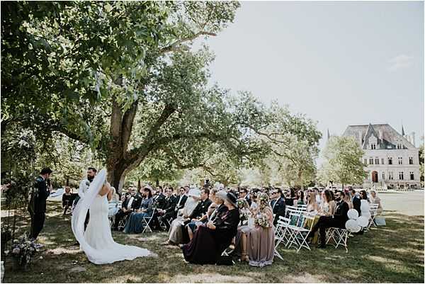 An outdoor wedding ceremony taking place on the grounds of a French chateau, visible in the background with its distinctive steep roof and dormer windows. The bride, wearing a long white dress with a flowing veil and train, stands facing the officiant and groom under a large mature tree, with a small floral arrangement on the ground nearby. Approximately 40–50 guests are seated in white folding chairs arranged in rows on the lawn, dressed in smart casual to formal attire, with some guests wearing hats. The image is a wide shot taken from behind and to the side of the ceremony, capturing both the couple and the full guest seating area in natural afternoon light. Potential venue feature image.