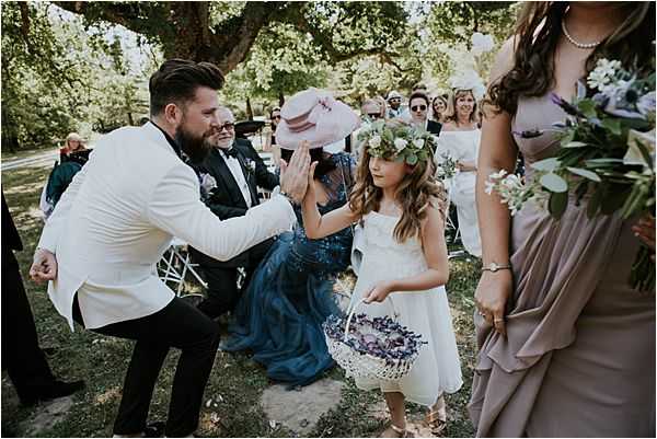 An outdoor wedding ceremony captures a candid moment between the groom and a flower girl exchanging a high-five along the aisle. The groom is wearing a white tuxedo jacket with black trousers and a small floral boutonniere. The flower girl is dressed in a white midi dress and wears a floral crown of white and greenery, holding a white wicker basket filled with purple flower petals. A bridesmaid in a taupe/mauve draped dress stands in the foreground right, holding a bouquet of white flowers and eucalyptus. Seated guests in the background include a woman in a teal/navy blue dress and another in a pale pink wide-brimmed hat. The ceremony takes place outdoors beneath large trees, with white folding chairs arranged along the aisle. The overall styling includes a soft, romantic palette of white, taupe, and purple with a garden-inspired aesthetic. Medium wide shot with a shallow depth of field.
