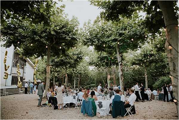 An outdoor wedding reception taking place in a tree-lined courtyard or garden with a gravel ground surface. Approximately 50-80 guests are seated at long rectangular tables draped with white linens, arranged beneath a canopy of tall mature trees strung with warm-toned fairy lights. A white timber-framed building is visible on the left side, decorated with large gold balloon letters. Guests are dressed in a mix of formal and smart-casual attire, including a woman in a teal/emerald gown prominently visible in the foreground. The overall styling is relaxed and festive, with a natural, informal outdoor reception atmosphere. Wide-angle shot capturing the full scene from a slightly elevated perspective.