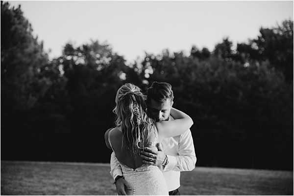 This black-and-white portrait shows a couple embracing outdoors on an open lawn, photographed from behind. The bride wears a lace or textured fitted gown with a low or open back, her hair styled in a loose half-up arrangement with soft waves falling down her back and what appears to be a decorative hair accessory. The groom, dressed in a light-colored suit jacket, holds her close with his face bowed toward her. The image is shot at a medium distance with soft, low contrast tones suggesting dusk or golden hour light, with dark tree lines forming the background.