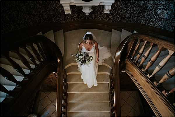 A bride descends a grand curved stone staircase inside a chateau or manor house, photographed from directly above in a dramatic overhead portrait composition. She wears a fitted ivory lace gown with thin straps and a cathedral-length veil, and carries a loose, organic bouquet featuring white blooms and trailing greenery with eucalyptus-style foliage. The interior features dark ornate wallpaper with a floral damask pattern, a carved white plaster ceiling detail, and a polished dark wood banister with turned spindles. The overhead angle highlights the sweeping spiral geometry of the staircase against the worn stone steps. Potential venue feature image.