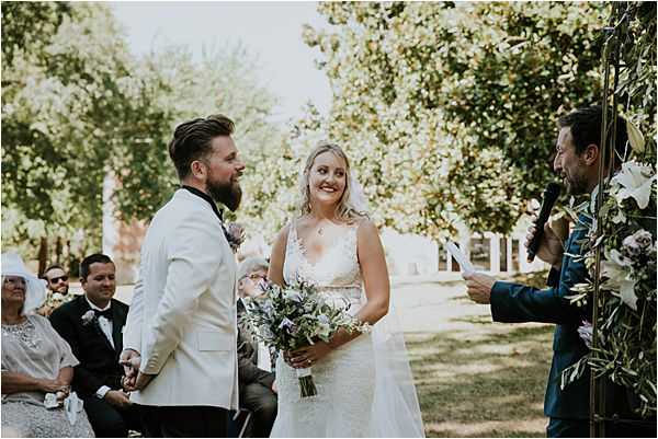 An outdoor wedding ceremony captured in a medium-wide shot, showing the bride and groom standing at the altar facing each other while an officiant in a navy suit reads from papers to their right. The bride wears a fitted lace gown with a V-neckline and holds a loose, garden-style bouquet of purple, lavender, and white blooms with trailing greenery. The groom is dressed in a white blazer with a black lapel. A gold arch decorated with olive branches and white and mauve florals frames the right side of the frame. Seated guests are visible in the background, including a woman in a wide-brimmed white hat, with the ceremony set in a shaded garden area with mature trees.