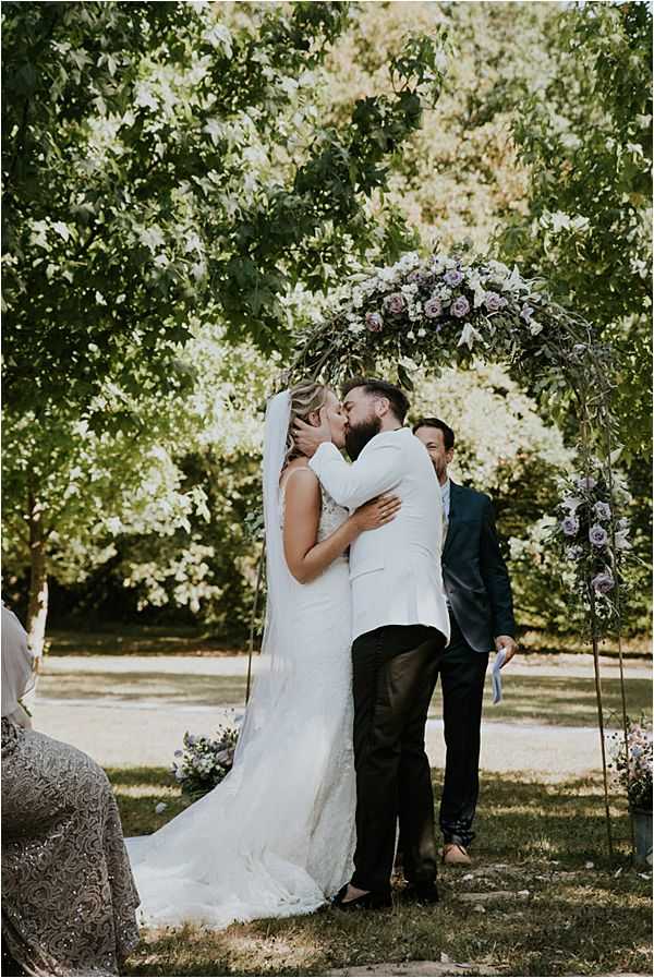 The bride and groom share their first kiss during an outdoor ceremony beneath a metal arch decorated with lavender and dusty mauve roses, white blooms, and trailing greenery. The bride wears a fitted lace white gown with a cathedral-length veil, while the groom is dressed in a white blazer with dark trousers. An officiant in a dark teal suit stands behind them holding ceremony notes, and a seated guest in a silver embroidered outfit is partially visible in the foreground. The setting is a shaded garden area with large leafy trees, and small floral arrangements in the same lavender and mauve palette are placed at the base of the arch. The shot is a medium full-length portrait taken from a slight distance, capturing the full arch and the couple's embrace.
