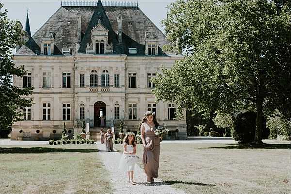 A bridesmaid in a taupe/mocha draped dress and a flower girl in a white dress walk together down the front path of a French chateau, moving toward the camera. Both wear floral crowns made of greenery and small white blooms, and the bridesmaid carries a loosely arranged bouquet in neutral and white tones. In the background, two additional bridesmaids in matching taupe dresses stand near the chateau's grand entrance steps. The wedding style is boho with a natural, earthy palette. The chateau is a classic French mansion with pale stone facade, ornate mansard roof with dark slate tiles, and symmetrical windows. Wide shot taken from ground level on the main driveway. Potential venue feature image.