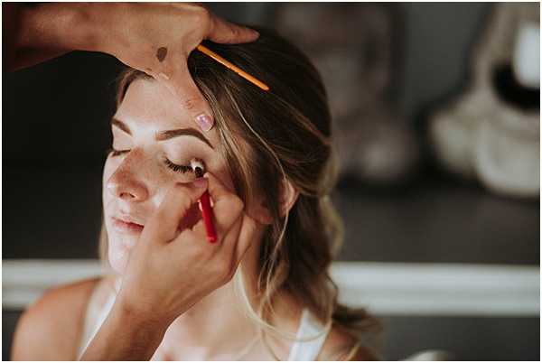 A close-up portrait of a bride during the getting-ready phase, with a makeup artist applying eyeliner using a red pencil while holding an orange makeup brush at the hairline. The bride has her eyes partially closed, wearing what appears to be a white bridal robe or top, with her blonde hair worn down in loose waves. The background is softly blurred with dark and neutral tones, suggesting an indoor setting with natural light coming from one side.