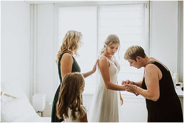 A getting-ready moment captured indoors in a bright, white room where a bride in a white sleeveless gown with a flowing skirt is being assisted by two women and a young girl. A woman in a dark green dress stands behind the bride with a hand on her back, while an older woman in a black sleeveless dress leans forward to fasten a bracelet or adjust jewelry on the bride's wrist. A young girl with brown hair stands in the foreground facing the bride. The room features white walls, white bedding, and sheer white curtains, creating a clean, minimal aesthetic. Medium portrait shot with natural window light from behind.